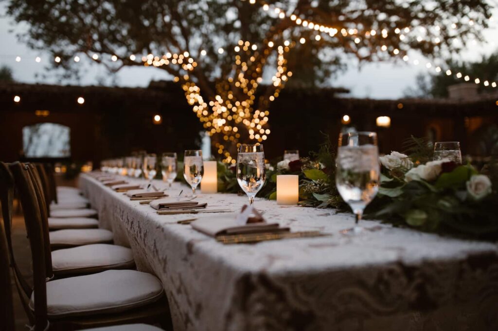 Long dinner table set beneath a tree wrapped in warm lights