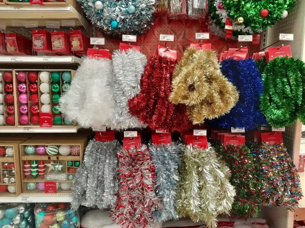 Store display featuring colorful Christmas decorations, including tinsel garlands in red, gold, silver, blue, and green, along with ornaments and wreaths arranged on shelves.