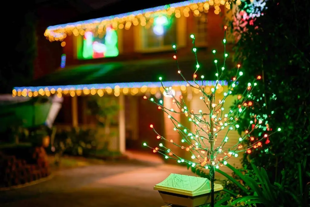 Small decorative tree covered in red and green LED lights in the foreground, with a house in the background adorned with yellow and blue Christmas lights along the roofline.