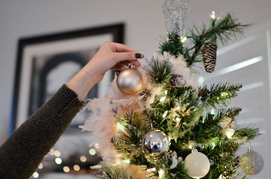 Close-up of a person’s hand hanging a gold ornament on a decorated Christmas tree adorned with white feathers, pinecones, and glowing string lights.