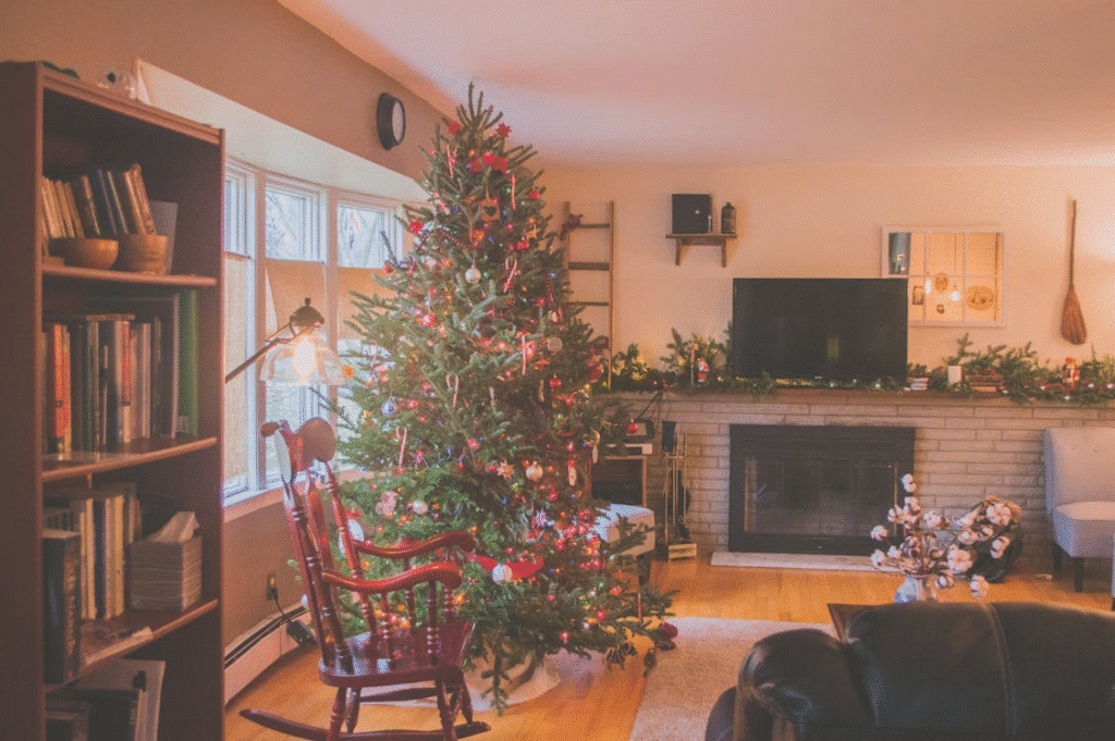 Cozy living room decorated for Christmas with a large tree adorned with ornaments and lights, a fireplace with garland, and a red rocking chair beside the window.