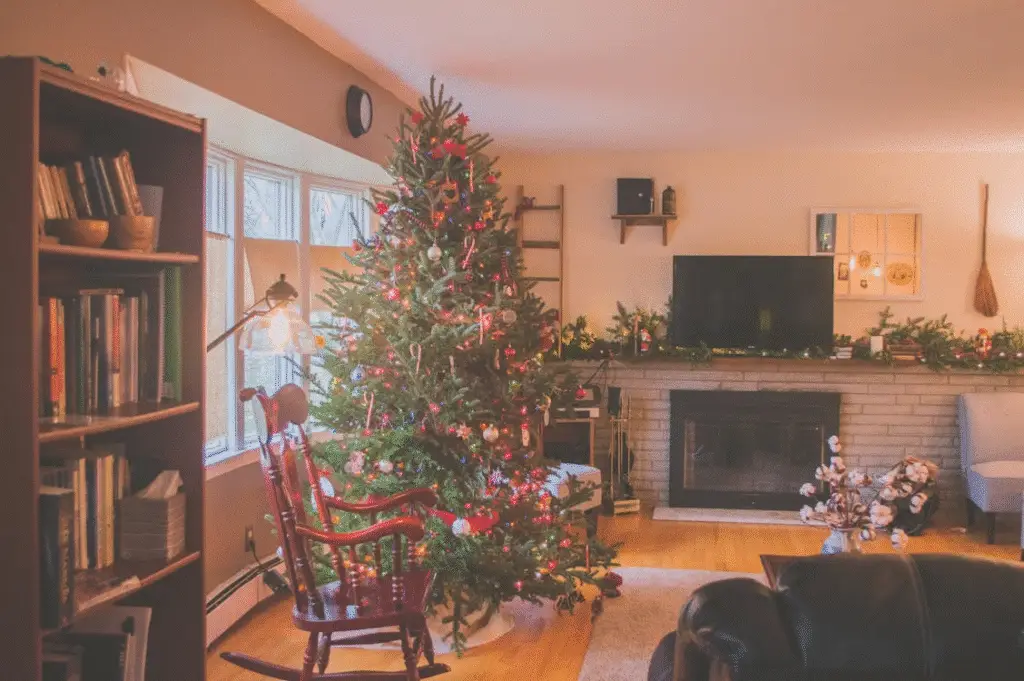 Cozy living room decorated for Christmas with a large tree adorned with ornaments and lights, a fireplace with garland, and a red rocking chair beside the window.