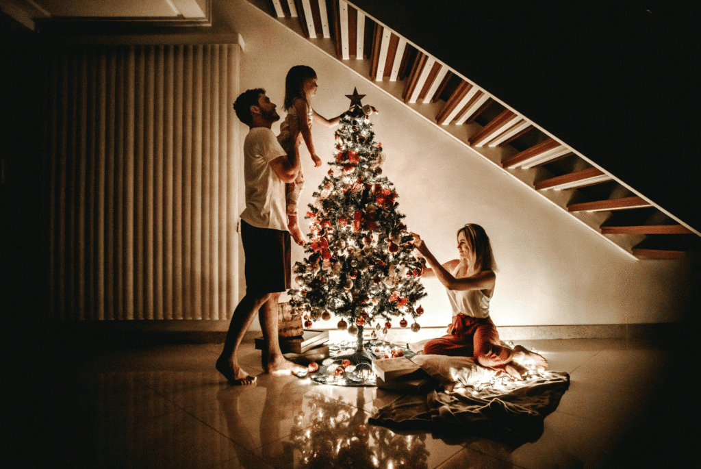 Family decorating a Christmas tree together in a warmly lit living room, with a father lifting his daughter to place an ornament while the mother arranges decorations near the base of the tree.