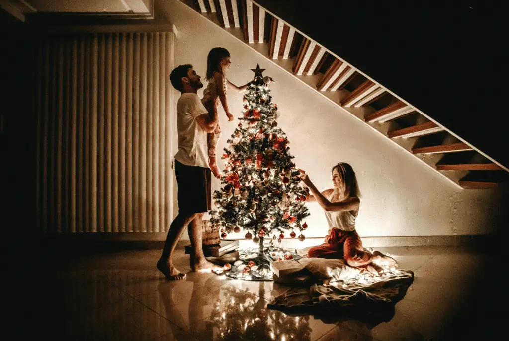 Family decorating a Christmas tree together in a warmly lit living room, with a father lifting his daughter to place an ornament while the mother arranges decorations near the base of the tree.