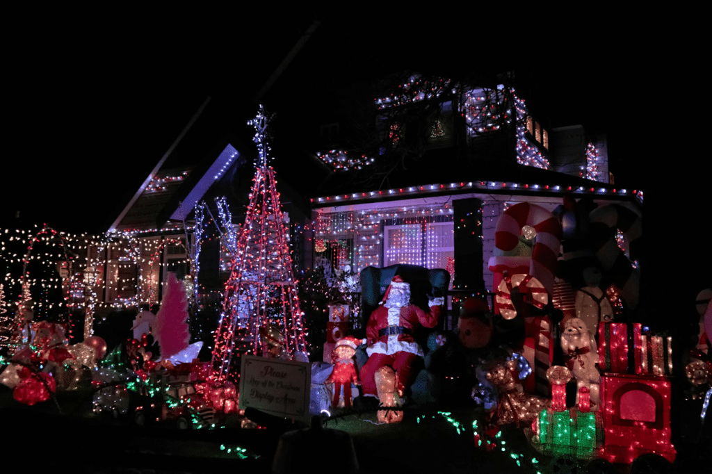 Elaborate Christmas light display covering a house and front yard, featuring multicolored string lights, light-up decorations, inflatables, a Santa figure, and festive props creating a vibrant holiday scene.