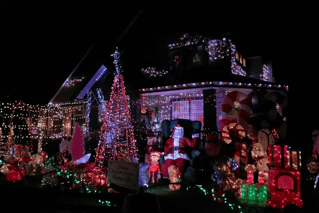 Elaborate Christmas light display covering a house and front yard, featuring multicolored string lights, light-up decorations, inflatables, a Santa figure, and festive props creating a vibrant holiday scene.