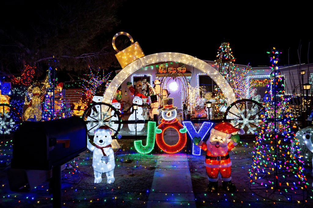 Extravagant Christmas light display featuring a giant illuminated ornament arch, colorful “JOY” sign with a snowman, Santa figure, polar bears, snowmen, and trees wrapped in multicolored lights, creating a vibrant and festive holiday scene.