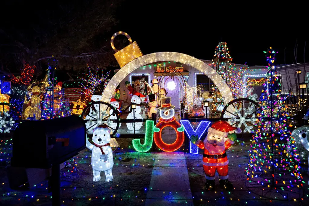 Extravagant Christmas light display featuring a giant illuminated ornament arch, colorful “JOY” sign with a snowman, Santa figure, polar bears, snowmen, and trees wrapped in multicolored lights, creating a vibrant and festive holiday scene.