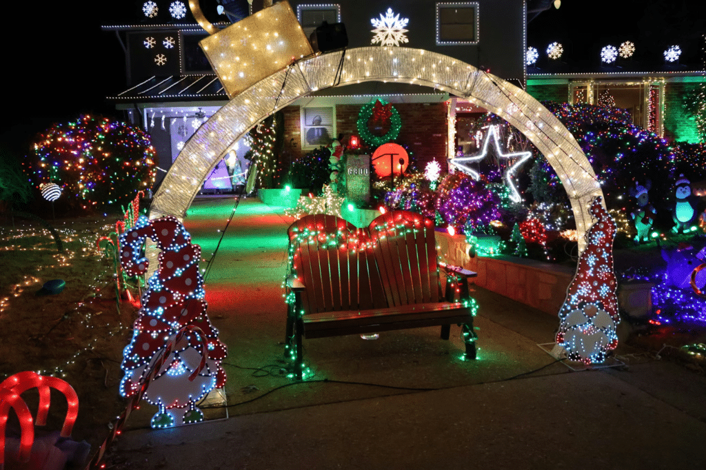 A festive front yard decorated with colorful Christmas lights, featuring a lighted archway with gnome figures, illuminated candy canes, a heart-shaped bench, and a house covered in twinkling lights, wreaths, and glowing holiday figures.