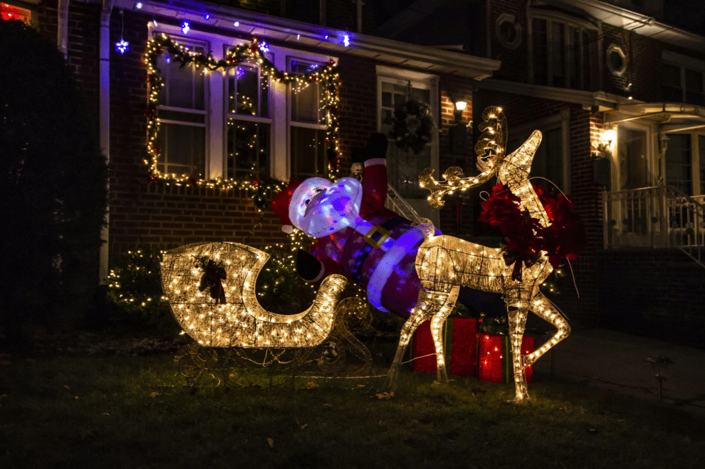 A festive front yard display featuring a glowing light-up reindeer pulling a sleigh filled with golden lights, with an inflatable Santa figure leaning inside, surrounded by twinkling garlands and Christmas lights decorating a brick house in the background.