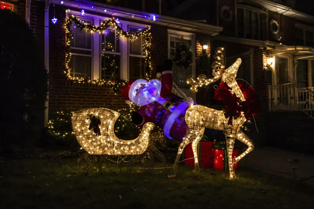 A festive front yard display featuring a glowing light-up reindeer pulling a sleigh filled with golden lights, with an inflatable Santa figure leaning inside, surrounded by twinkling garlands and Christmas lights decorating a brick house in the background.