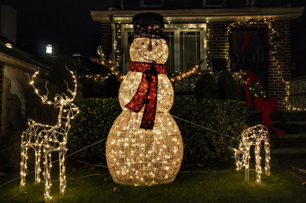 A glowing outdoor Christmas display featuring a large light-up snowman with a red scarf and top hat, flanked by two illuminated reindeer figures, set against a brick house decorated with white string lights, wreaths, and festive red bows.