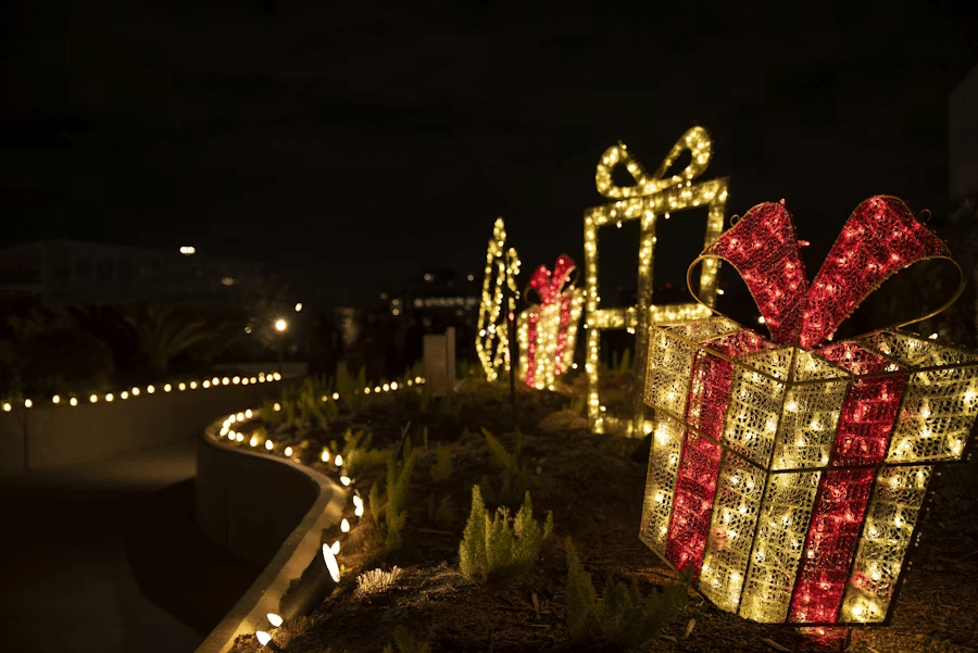 Outdoor holiday display featuring large illuminated Christmas presents wrapped in red and gold lights, with a pathway lined by warm white string lights at night.