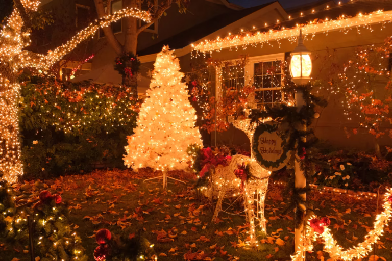 Festive front yard Christmas display featuring a glowing white artificial tree, light-wrapped trees and bushes, icicle lights on the roofline, and a lit reindeer figure with a “Happy Holidays” sign.